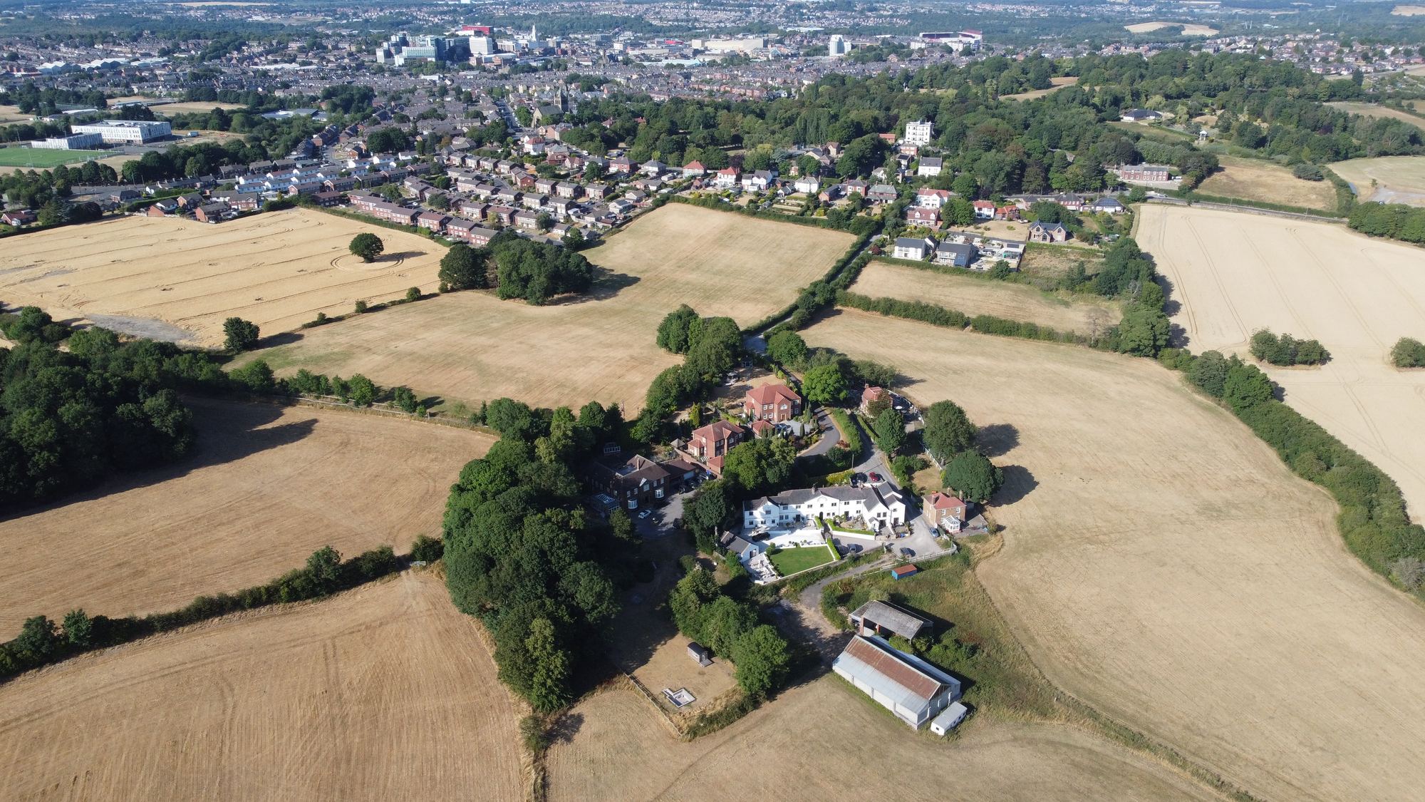 Aerial photograph looking north across open harvested farmland on the southern edge of Barnsley. A group of older buildings sits among mature trees in the middle distance, with hedged fields around them, the houses and schools of Kingstone beyond, and Barnsley town centre on the skyline.